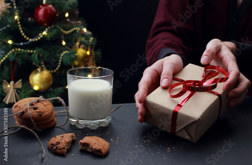 young european woman holds new year gift with red ribbon. A glass of milk and cookies with chocolate and crumbs for Santa on the background of a decorated Christmas tree, dark mood