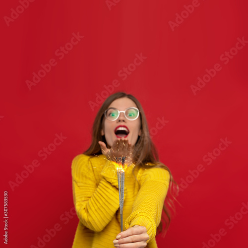 happy girl wears glasses and yellow jumper holding sparklers and blowing kiss, positive emotions and feelings, isolated on red background. Christmas concept