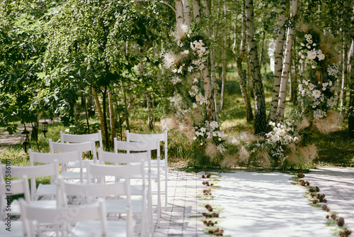 Place for wedding ceremony in garden, copy space. Wedding arch decorated with flowers and white chairs on each side of archway outdoors. Wedding setting