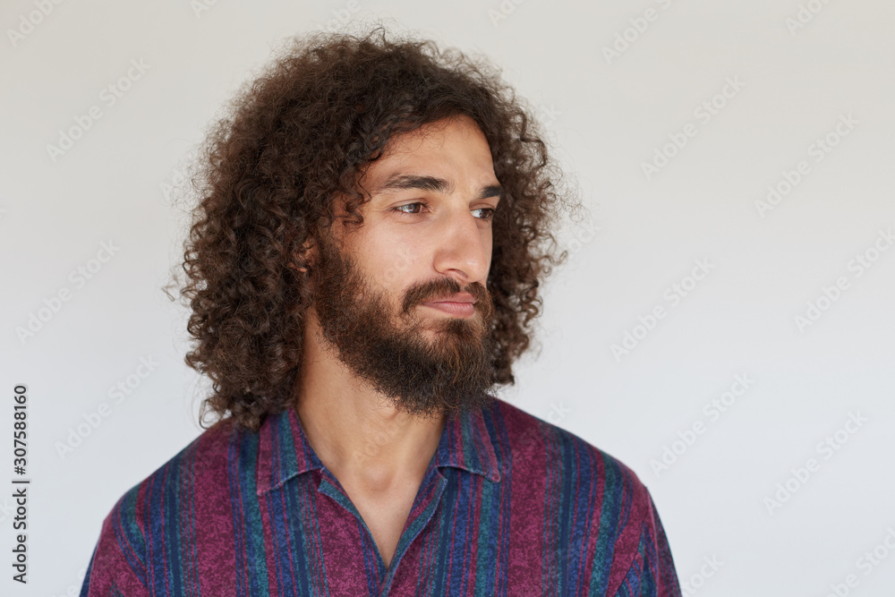 Portrait of pretty young brunette curly man with beard keeping lips folded while looking aside with calm face, wearing striped multi-colored shirt while posing against white background Portrait of pretty young brunette curly man with beard keeping lips folded while looking aside with calm face, wearing striped multi-colored shirt while posing against white background