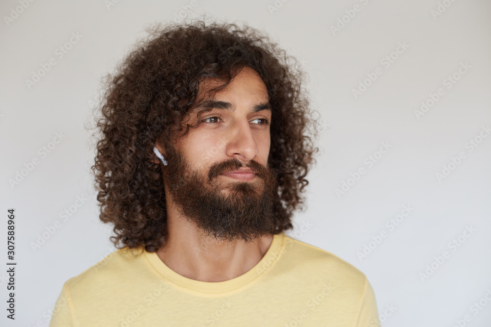 Handsome young bearded man with brown curly hair looking aside thoughtfully while listening to training with earphones, dressed in casual clothes while posing over white background Handsome young bearded man with brown curly hair looking aside thoughtfully while listening to training with earphones, dressed in casual clothes while posing over white background