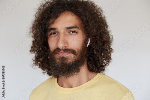 Close-up of beautiful young dark haired curly man with charming brown eyes looking positively to camera with light smile, wearing lush beard and listening to music in his headphones