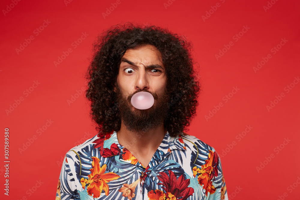 Indoor shot of funny brunette curly male with beard inflating ball with chewing gum while looking at camera with raised eyebrow, isolated against red background in casual clothes Indoor shot of funny brunette curly male with beard inflating ball with chewing gum while looking at camera with raised eyebrow, isolated against red background in casual clothes