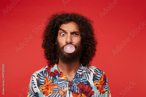 Fotografie Indoor shot of funny brunette curly male with beard inflating ball with chewing