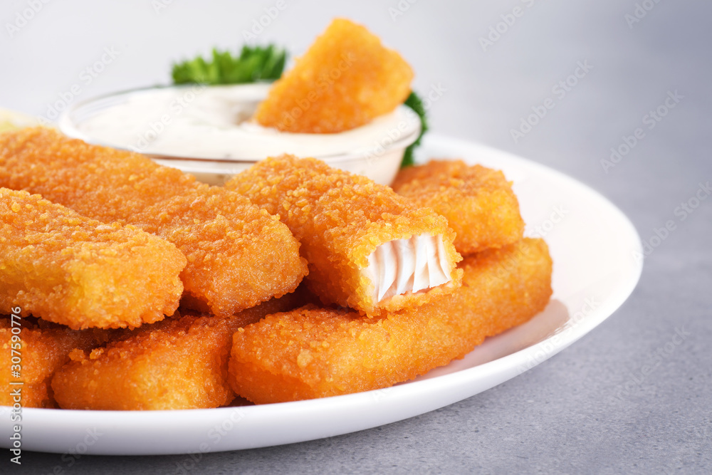 Fish fingers sticks on a plate with parsley, lemon and tartar sauce on a gray texture.