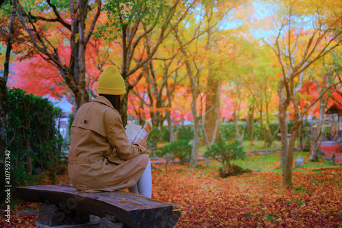 woman sitting on wooden chair under shadow of the autumn tree in public park, comfort reading a book in the light of morning of autumn season change