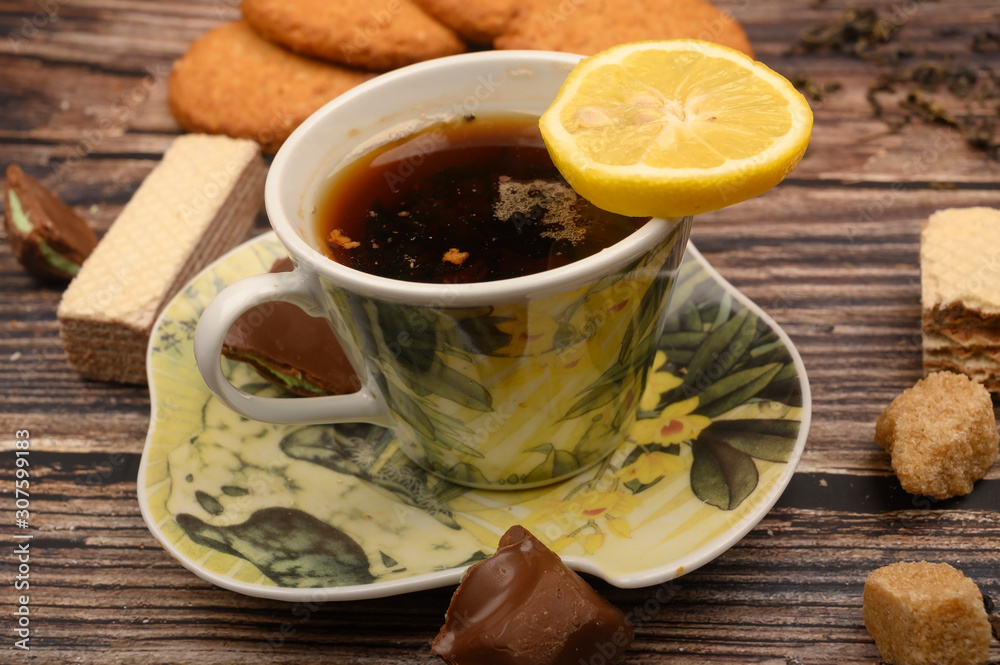 A Cup of black tea, tea leaves, pieces of brown sugar, oatmeal cookies, waffles, pieces of chocolate, lemon on a wooden background. Close up. A Cup of black tea, tea leaves, pieces of brown sugar, oatmeal cookies, waffles, pieces of chocolate, lemon on a wooden background. Close up.