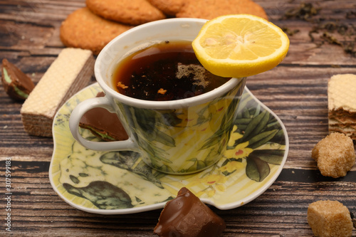 A Cup of black tea, tea leaves, pieces of brown sugar, oatmeal cookies, waffles, pieces of chocolate, lemon on a wooden background. Close up.