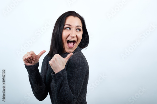Cheerful caucasian young woman over isolated white background in fashion black sweater pointing thumbs up with opened mouth, looking aside. Lifestyle concept