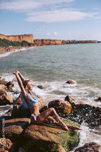 Luxury girl posing on a rocky seashore in a blue swimsuit. Spray water on the rocks.