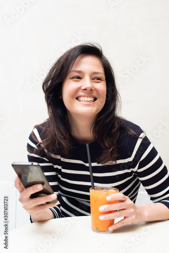happy young woman sitting at table with mobile phone and drink in hand