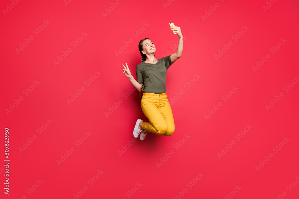 Full length photo of active lady jumping high making selfies showing v-sign symbol cheerful mood wear casual yellow pants green t-shirt isolated red color background Full length photo of active lady jumping high making selfies showing v-sign symbol cheerful mood wear casual yellow pants green t-shirt isolated red color background