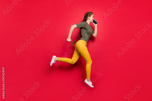 I'm on my way. Full length photo of active lady jumping high running fast meeting with friends speaking telephone wear casual pants t-shirt isolated red color background