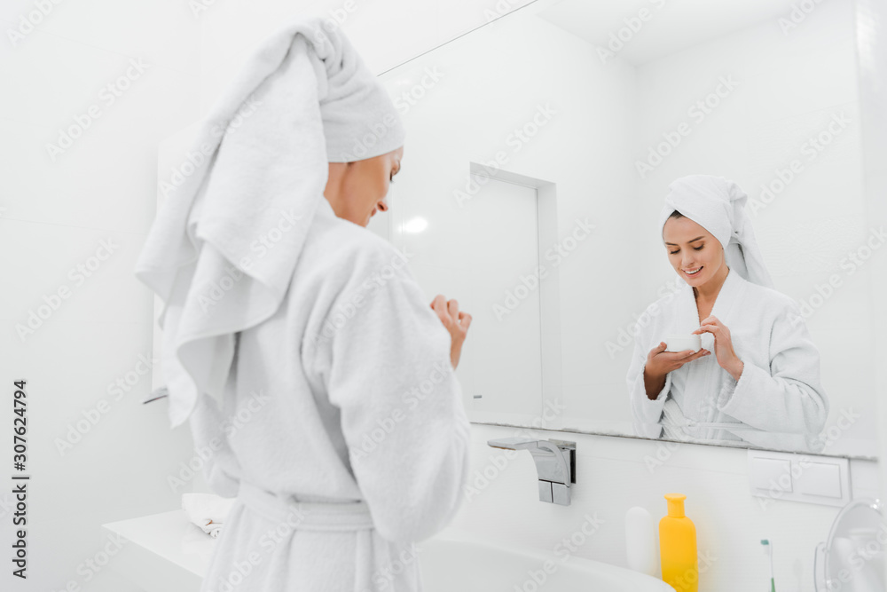 selective focus of happy woman holding container with cosmetic cream in bathroom selective focus of happy woman holding container with cosmetic cream in bathroom
