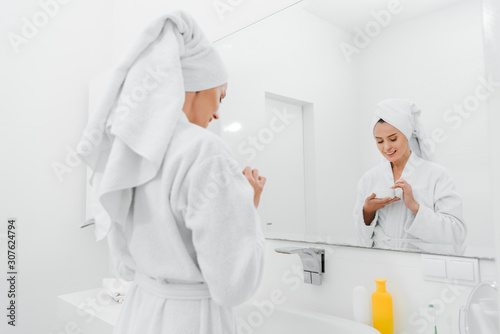 selective focus of happy woman holding container with cosmetic cream in bathroom