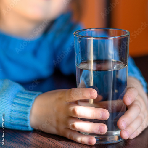 Little kid holding glass of water in kitchen. Glass is focused. Health and beauty concept