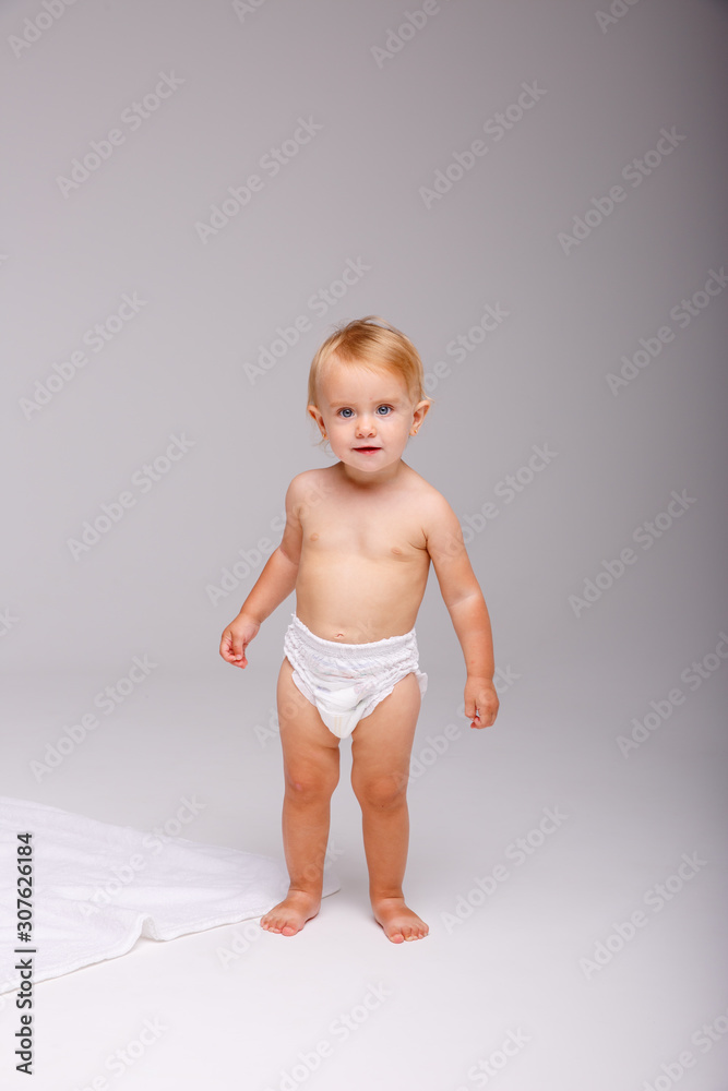 A baby girl in a diaper stands tall on a white background looking at the camera A baby girl in a diaper stands tall on a white background looking at the camera
