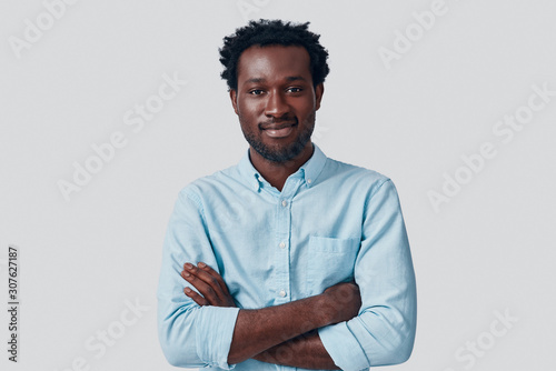 Handsome young African man looking at camera and keeping arms crossed while standing against grey background