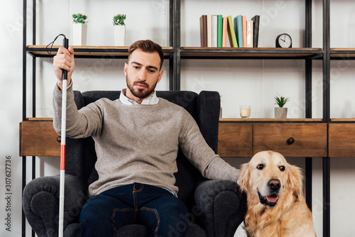 Blind man with walking stick sitting in armchair beside golden retriever at home photo