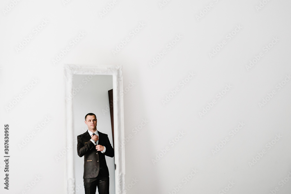 Fototapeta premium The man in a suit looks at his reflection in the mirror and straightens tie on shirt at home. clothing concept. preparing for a business meeting.