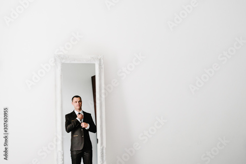 The man in a suit looks at his reflection in the mirror and straightens tie on shirt at home. clothing concept. preparing for a business meeting. © Serhii