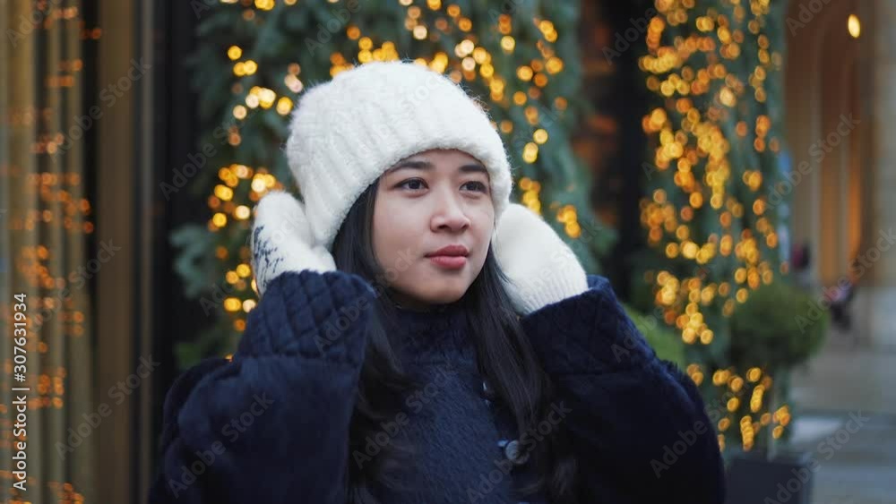 Pretty girl in Europe is outside near Christmas decorations. European street decorated for Christmas. Girl touch her white hat, she wears white mittens. Christmas tree lights and garlands on a shop. Pretty girl in Europe is outside near Christmas decorations. European street decorated for Christmas. Girl touch her white hat, she wears white mittens. Christmas tree lights and garlands on a shop.