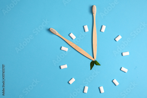 Two wooden bamboo eco friendly toothbrushes, green leaf and chewing gum in the form of hours on a blue background. Eco friendly and reuse concept. Flat lay, top view, copy space