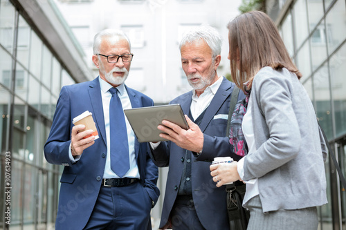 Small mixed group of businessmen and businesswoman meeting in front of a glass facade office building during coffee break, looking at sale reports on a tablet
