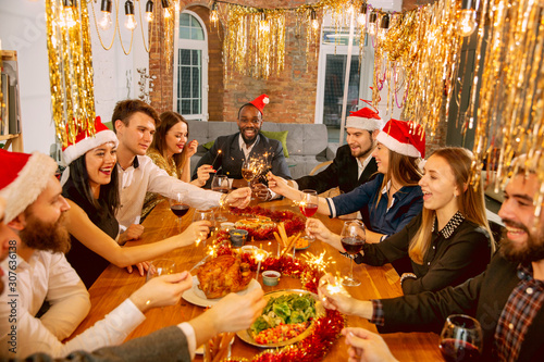 Happy co-workers celebrating while company New Year and Christmas party. Young caucasian people in celebrate attire with sparklers talk  drink wine  cheers. Concept of office culture  teamwork  winter