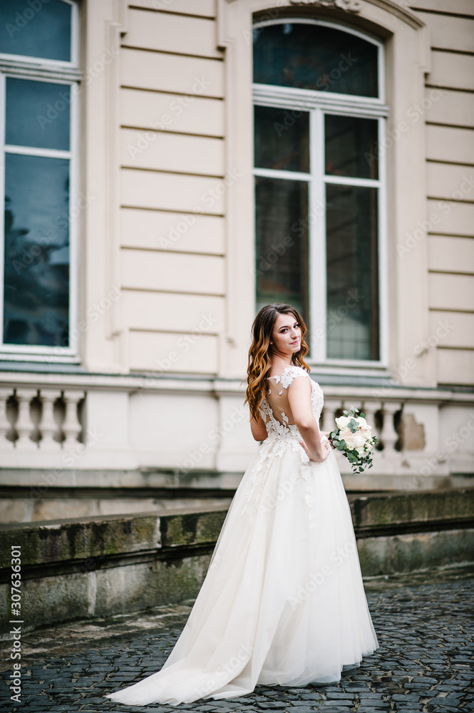 Fototapeta premium Portrait bride in wedding dress with a bouquet of flowers standing back near old building outside, vintage palace outdoor.