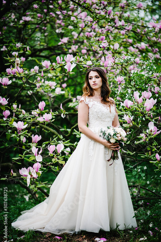 Beautiful bride in white dress with a wedding bouquet posing and standing on background nature of purple, pink flowers of magnolia and greens. outdoors.