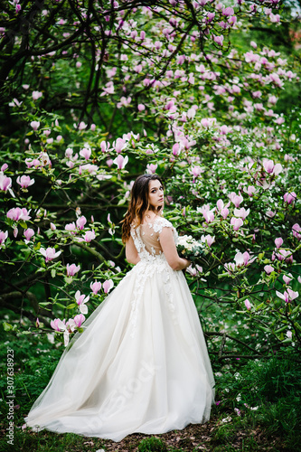 Glamorous bride with a wedding bouquet standing back, on background flowers of magnolia and greens. outdoors. Wedding location on the ceremony.