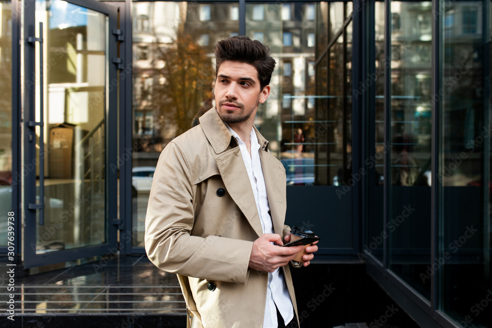 Young confident casual man in trench coat with cellphone intently looking  away on street Photos | Adobe Stock
