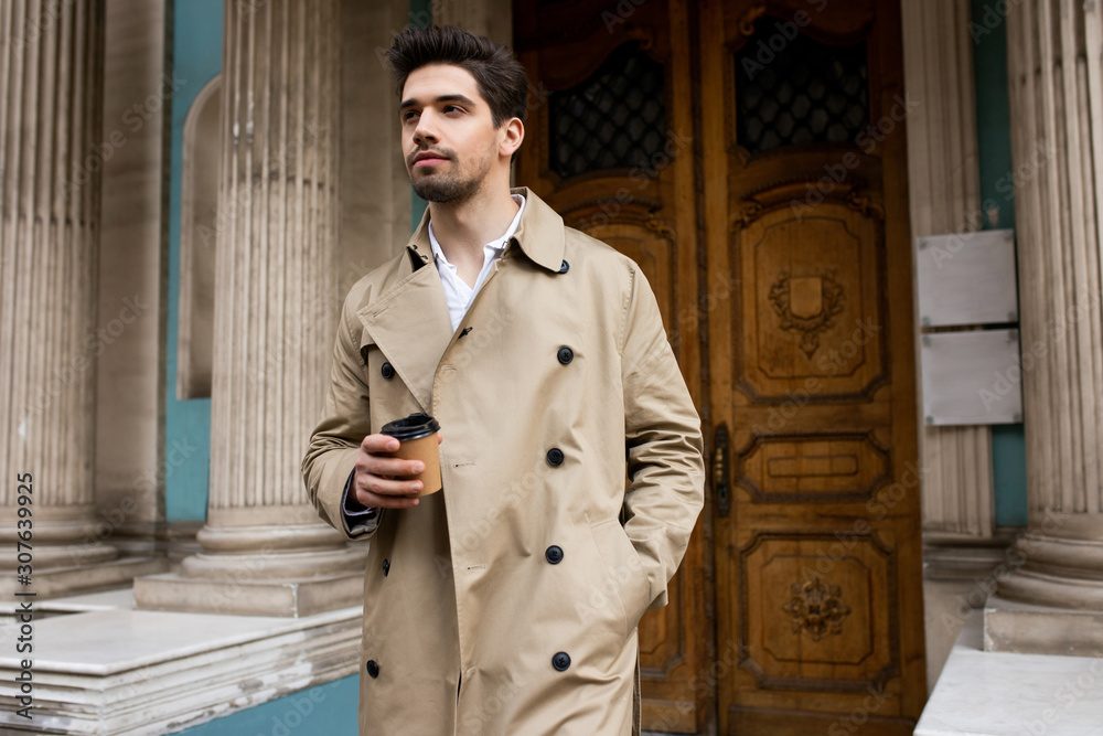 Young confident man in trench coat walking through city street with coffee  to go Photos | Adobe Stock