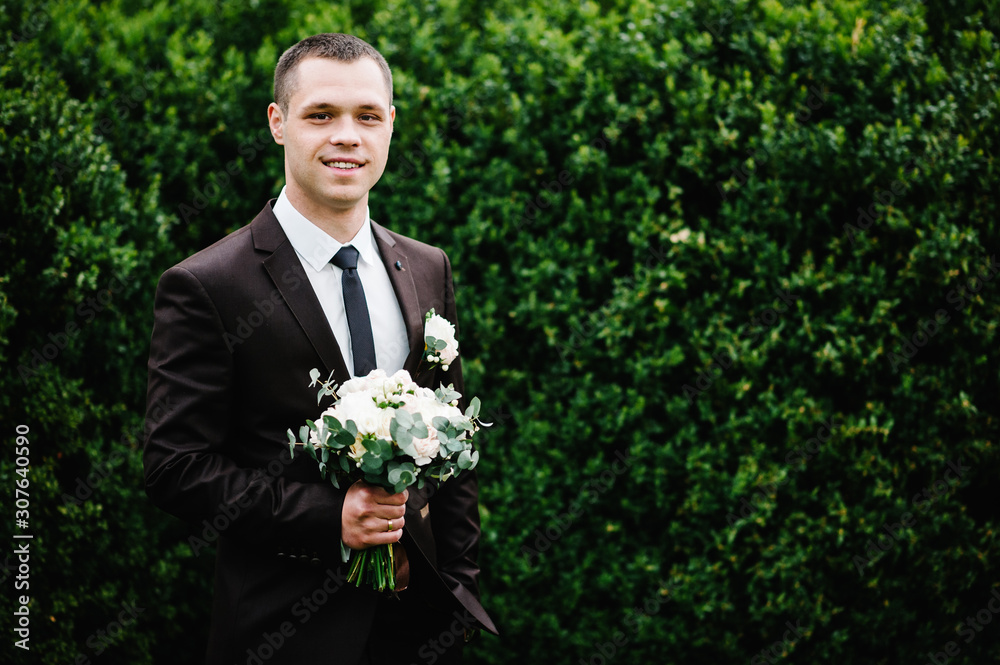 The attractive groom in a suit and tie with wedding bouquet flowers, and a boutonniere or buttonhole on the jacket is standing on the background of greenery in park. The attractive groom in a suit and tie with wedding bouquet flowers, and a boutonniere or buttonhole on the jacket is standing on the background of greenery in park.