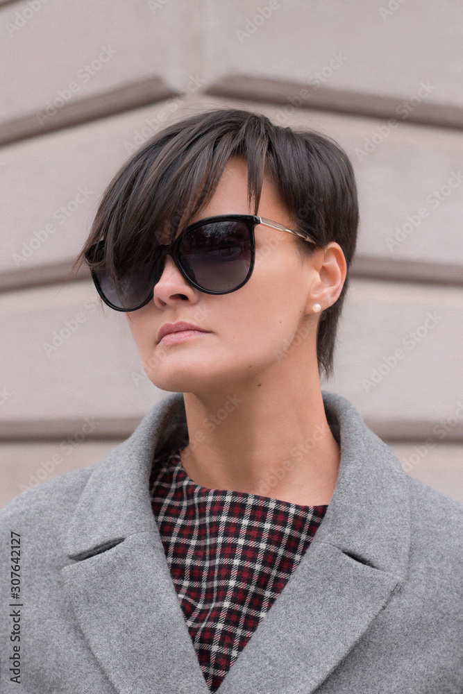 Autumn fashion outdoor. The brunette girl with short hair in fashionable stylish grey coat and sunglasses, posing on the background of building. Street fashion.Autumnal lifestyle