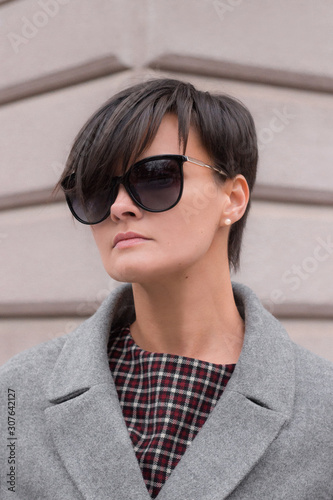Obraz na płótnie Autumn fashion outdoor. The brunette girl with short hair in fashionable stylish grey coat and sunglasses, posing on the background of building. Street fashion.Autumnal lifestyle w salonie