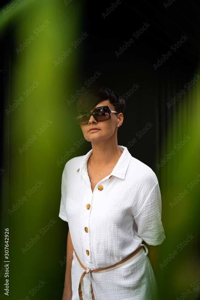 Beautiful woman with short brunette hair in white clothes and sunglasses. Fashion street photography. Fashion model posing on dark background.