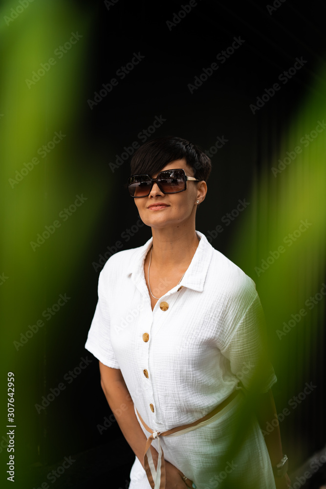 Beautiful woman with short brunette hair in white clothes and sunglasses. Fashion street photography. Fashion model posing on dark background.