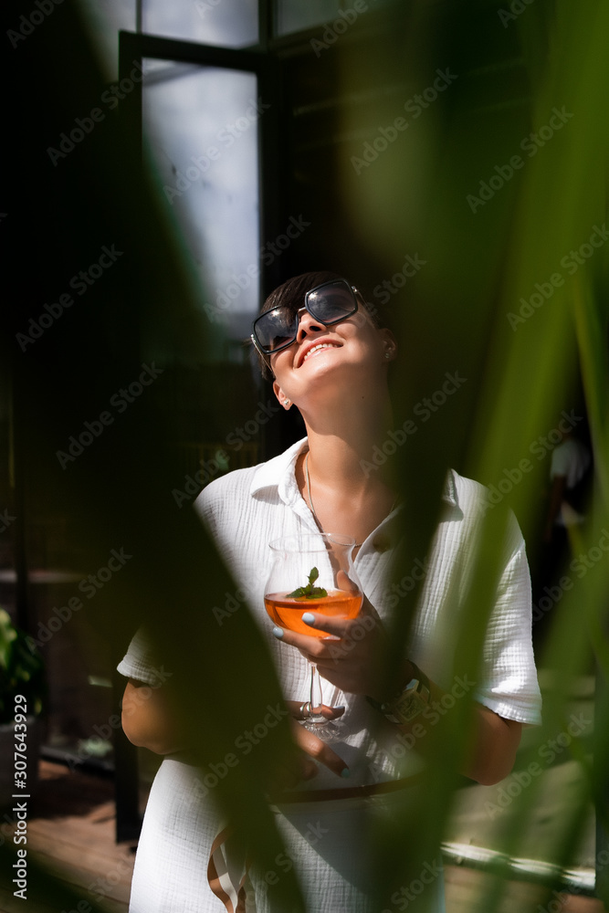 Beautiful woman with short brunette hair in white clothes and sunglasses. Fashion street photography. Fashion model posing with aperol cocktail on wood background.
