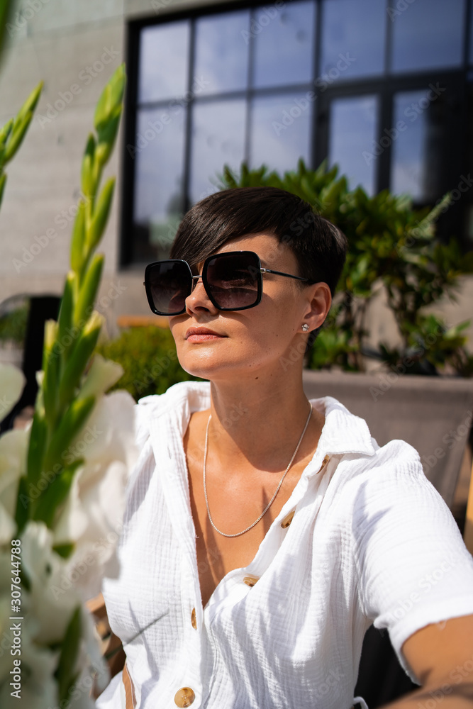 Beautiful woman with short brunette hair in white clothes and sunglasses. Fashion photography. Fashion model posing with flowers on wood background.