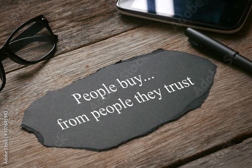 Glasses,mobile phone,pen,a a piece of black paper written with People buy...from people they trust on wooden background. photo