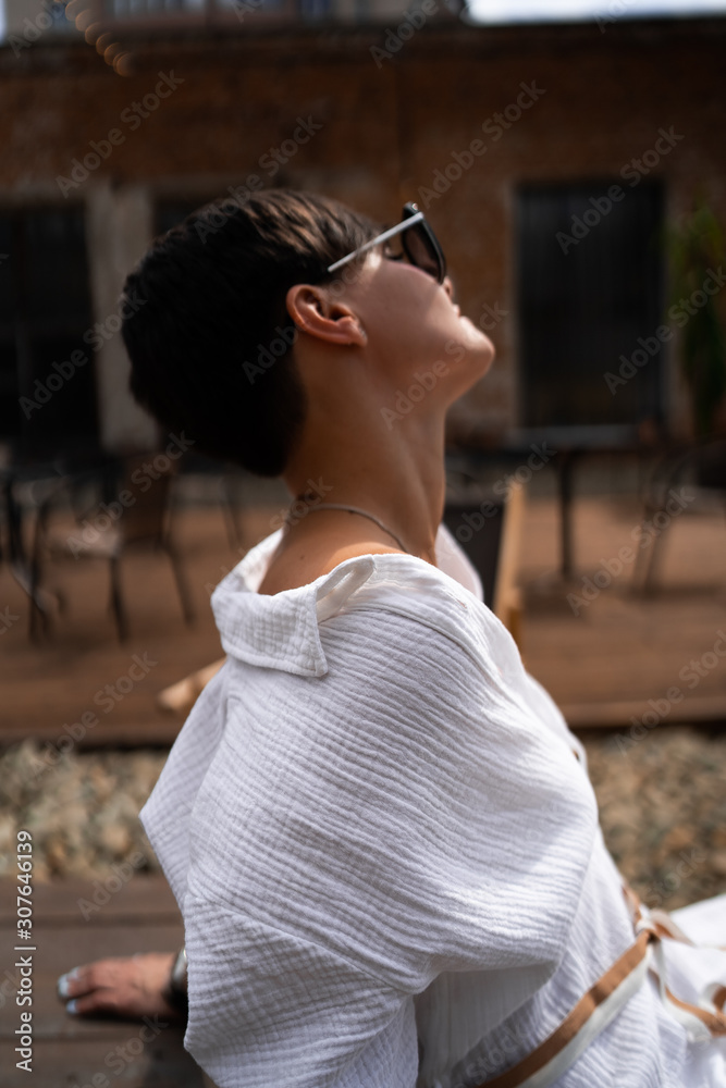 Beautiful woman with short brunette hair in white clothes and sunglasses. Fashion street photography. Fashion model is sitting and relax on brown wood background. sunglasses shop