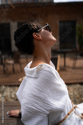 Obraz na płótnie Beautiful woman with short brunette hair in white clothes and sunglasses. Fashion street photography. Fashion model is sitting and relax on brown wood background. sunglasses shop w salonie
