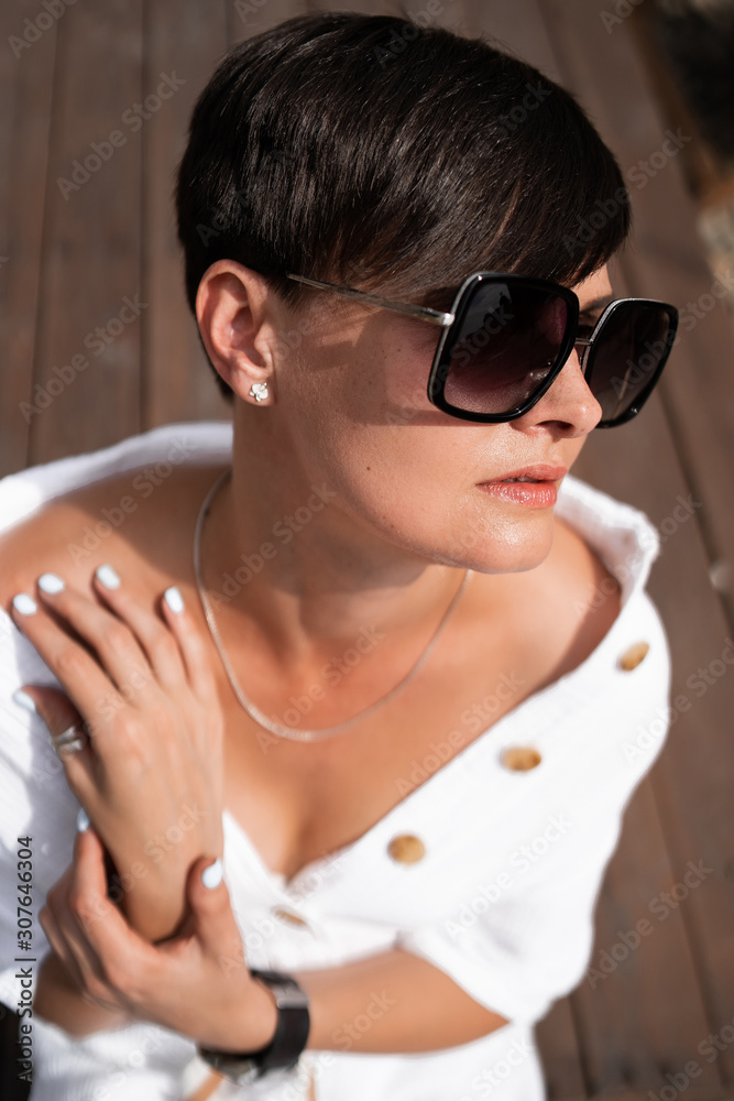 Beautiful woman with short brunette hair in white clothes and sunglasses. Fashion street photography. Fashion model is sitting and relax on brown wood background. sunglasses shop