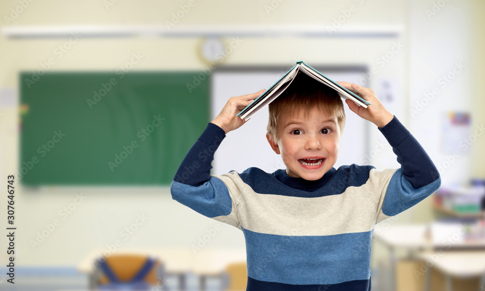 Fototapeta premium school, education and learning concept - smiling little boy with roof of book on top of his head over classroom background