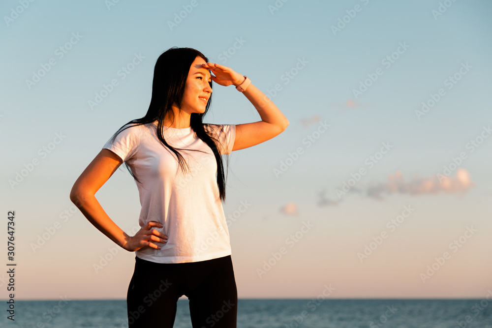 The concept of sport and a healthy lifestyle. A young woman in sports clothes , standing on the coastal rocks, resting her hand on her hips and looking into the distance. Horizontal and copy space