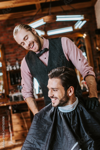 handsome barber in latex gloves putting hand on shoulder of happy man in barbershop