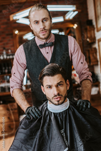 handsome barber in latex gloves putting hands on shoulders of man in barbershop