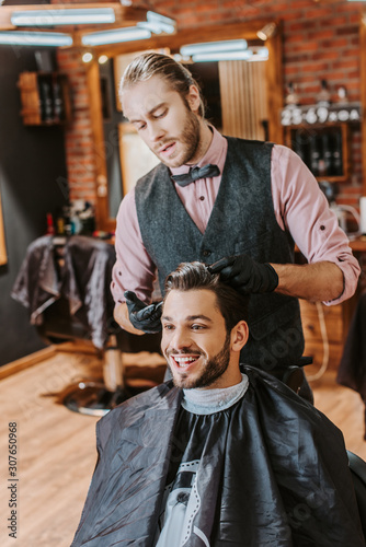 handsome barber in black latex gloves touching hair of happy man in barbershop